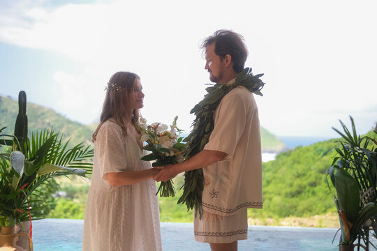 Outdoor wedding ceremony with ocean and green hills in the background. Bride in white dress and groom standing in the center in the arch of plants and holding hands. Saying wedding vows. Love. - Powered by Adobe