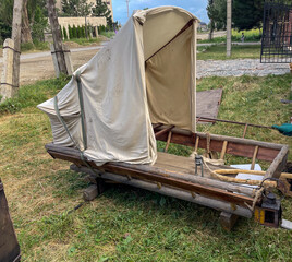 A rustic wooden cart features a white fabric canopy, resting on a grassy area. The cart has wooden...