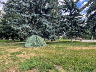 A green camping tent is pitched on grassy ground, surrounded by tall evergreen trees. The sky is...