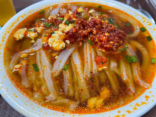 A close-up of a steaming bowl of noodles in a rich, red broth, topped with minced garlic, green onions, and a dollop of spicy chili paste, showcasing a hearty meal.