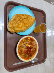 A bowl of spicy noodle soup with red chili flakes, garnished with green onions and egg, accompanied by a golden fried bread and a glass of yellow beverage.