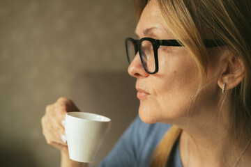 Happy dreamy middle aged woman is staying in living room with cup of black tea or coffee, looking away, Peaceful mature lady enjoying no stress calm positive pastime alone at home