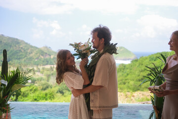 Outdoor wedding ceremony with ocean and green hills in the background. Bride in white dress and...