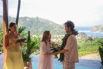 Outdoor wedding ceremony with ocean and green hills in the background. Bride in white dress and...