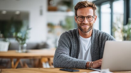 Confident young man with glasses working remotely on a laptop in a bright, modern home office, wearing a casual sweater and smiling, surrounded by natural light and minimalist decor