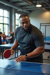 A plus-size black man is playing table tennis in a modern gym with large windows and exercise equipment. The atmosphere is energetic and competitive