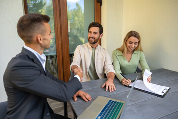 Real estate agent finalizing property contract with couple at outdoor table with laptop and...