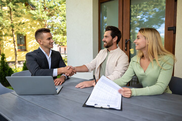Real estate agent signing agreement with couple at outdoor table with laptop and documents for...