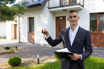 Real estate agent holding keys and clipboard in front of modern house for property