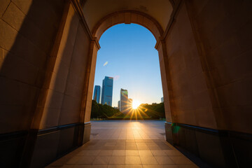 View of skyscrapers through stone archway at sunset with sunburst and blue sky in the background