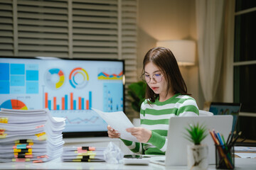 A stressed woman works late at a cluttered desk, surrounded by papers and charts, overwhelmed with...