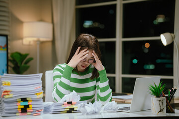 A stressed woman works late at a cluttered desk, surrounded by papers and charts, overwhelmed with...