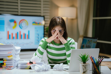 A stressed woman works late at a cluttered desk, surrounded by papers and charts, overwhelmed with...