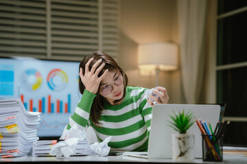A stressed woman works late at a cluttered desk, surrounded by papers and charts, overwhelmed with...