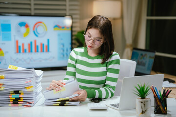 A focused woman works at night, analyzing financial charts and data. She smiles while reviewing...