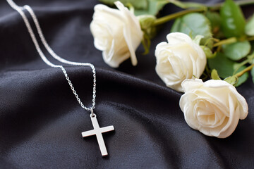 Elegant cross necklace with white roses, funeral flower bouquet on black silk fabric background