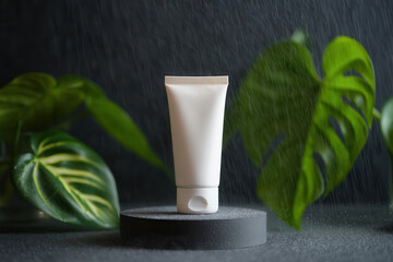 A white cosmetic tube on a gray pedestal surrounded by green leaves against a dark background