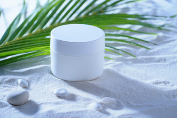 White cosmetic jar on white sand with palm leaf and small stones in a bright studio setting