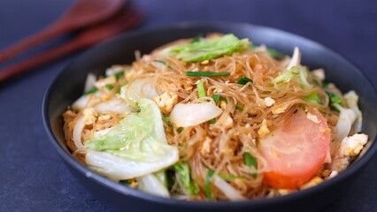 Stir-fried glass noodle with egg and vegetables in black bowl.