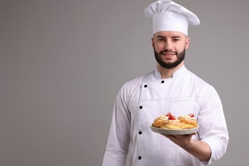 Happy confectioner in uniform holding delicious profiteroles with strawberries on light grey background