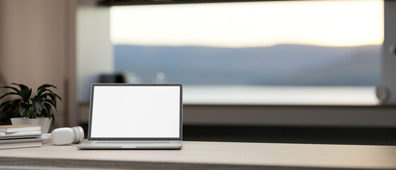 White screen laptop with books and headphones on wooden table in a room with scenery outside window.