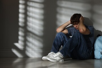 Teen problems. Upset teenage boy with backpack near light wall indoors, space for text