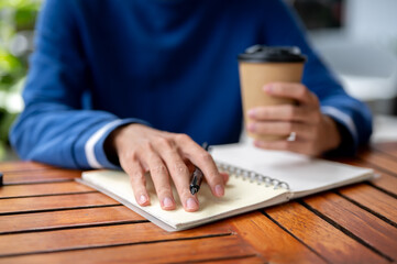Close up of hand with pen and other holding coffee over notebook at wood table in cafe or canteen.