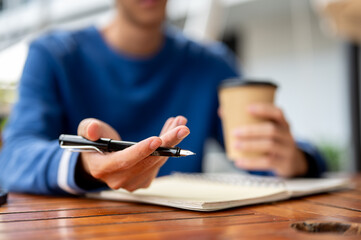 Close up of hands holding pen open palm and coffee over notebook at wooden table in cafe or canteen.