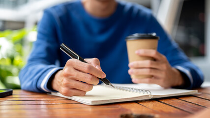 Close up of hands holding pen writing and coffee over notebook at wooden table in a cafe or canteen.