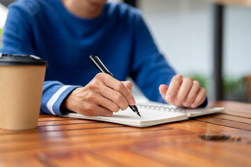 Close up of a hand holding pen writing in notebook aside coffee at wooden table in cafe or canteen.