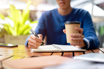 Close up of hands holding coffee and pen writing in notebook at wooden table in a cafe or canteen.