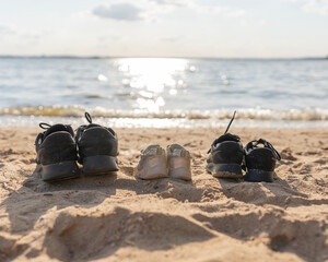 Three pairs of shoes — two adult and one child — placed on a sandy beach near the shoreline. Family footwear symbolizing vacation, togetherness, and summer relaxation by the sea on a sunny day.