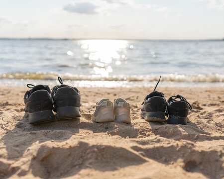 Three pairs of shoes — two adult and one child — placed on a sandy beach near the shoreline. Family footwear symbolizing vacation, togetherness, and summer relaxation by the sea on a sunny day. 