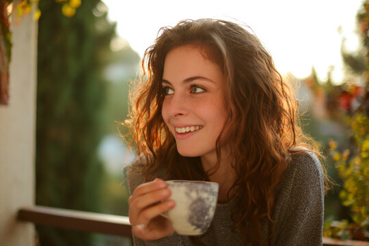 young woman with wavy brown hair enjoys warm drink on balcony, smiling brightly in soft sunlight. scene conveys sense of warmth foliage weather season and happiness