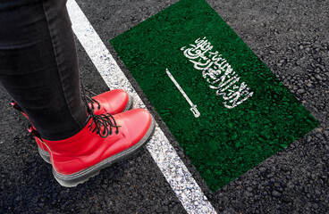 a woman with a boots standing on asphalt next to flag of Saudi Arabia and border
