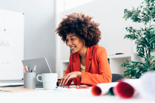 Happy businesswoman with Afro hairstyle using laptop at desk in office