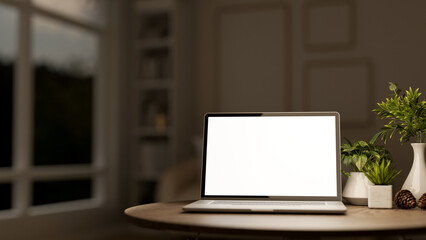 White screen laptop and acorn with vase and green pot plants on wooden table in a dim lit living room.