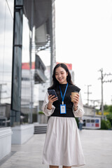 Smiling office worker or event staff holding coffee looking at phone while walking outside building