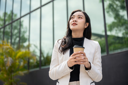 Asian office worker or businesswoman holding coffee with both hand while standing outside building.
