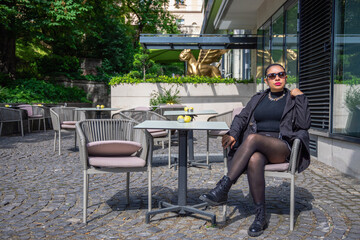 A confident woman in a black outfit poses stylishly at an outdoor cafe.