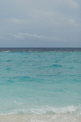 Minimalist seascape with gentle waves lapping the sandy shore under a cloudy sky. Soft turquoise hues and calm ocean view create a peaceful tropical atmosphere.