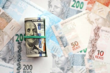 Roll of US dollar bills lies on big amount of Brazilian reais banknotes close up