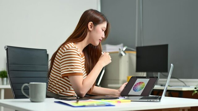 Young graphic designer using pen tablet and laptop at creative desk in modern office. Surrounded by color palettes