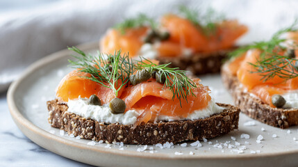 A close-up of open-faced sandwiches on a plate. The sandwiches feature dark rye bread topped with cream cheese, smoked salmon, capers, and fresh dill.