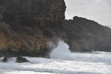 Waves crash against the rocky cliffs of Praia da Adraga, in Sintra-Cascais Natural Park, Portugal, under a cloudy sky. A dramatic seascape of raw Atlantic beauty.