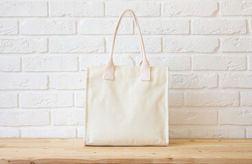 Cream color blank canvas tote bag mockup on wooden table top with white brick wall as background