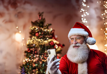 Weihnachtsmann mit wei&szlig;em Bart und Brille mit Smartphone vor Weihnachtsbaum