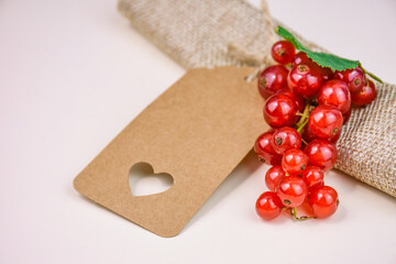 Blank kraft paper tag with cut out heart lying next to bunch of fresh red currant berries and green leaf on soft neutral background.