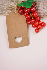 Blank kraft paper tag with cut out heart lying next to bunch of fresh red currant berries and green leaf on soft neutral background.