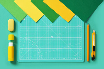Top view of a craft table in green and yellow colors. Craft knife, pencil, eraser, glue, cutting mat, colored paper. Space for your text.